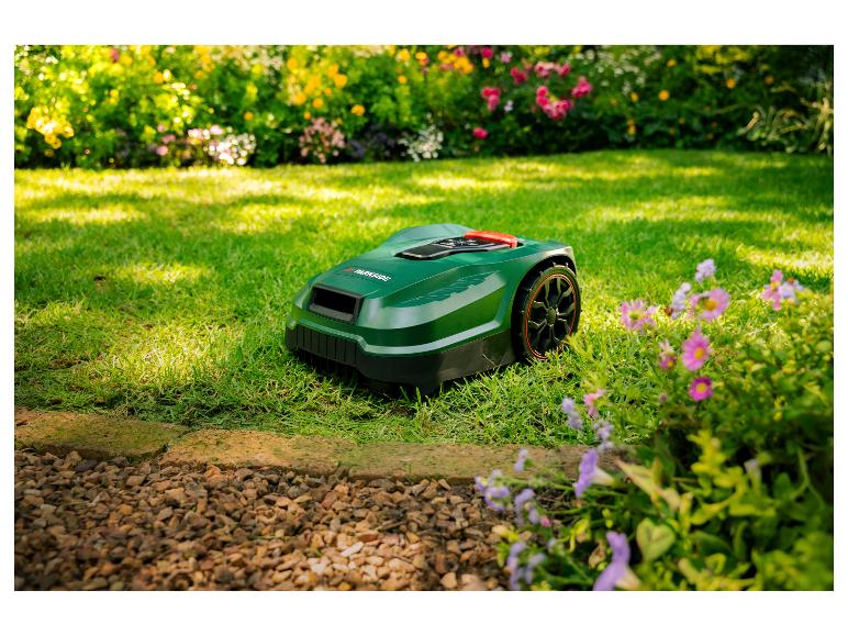 Een groene robotmaaier op een gazon met bloemen en struiken op de achtergrond.