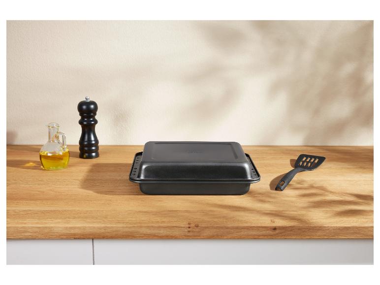 Zwarte braadslede met deksel, een oliefles en een pepermolen op een houten tafel.