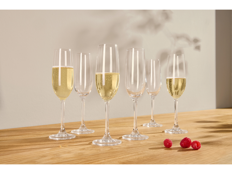 Zes champagneglazen, sommige gevuld met mousserende wijn, en drie frambozen op een houten tafel.