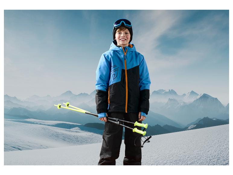 Een jongen in een blauwe jas en zwarte broek houdt ski-stokken vast in een besneeuwd landschap.