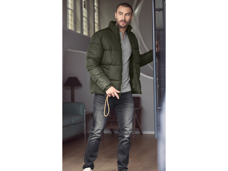 Man in olijfgroene pufferjas, grijs T-shirt en donkere jeans met een riem.