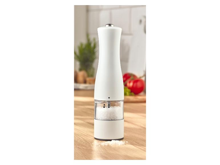 Witte zoutmolen met zichtbaar zeezout op een houten tafel met groenten op de achtergrond.