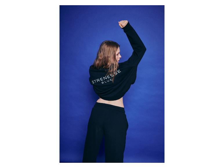 Vrouw in zwarte hoodie en joggingbroek, met opgeheven arm, tegen blauwe achtergrond.