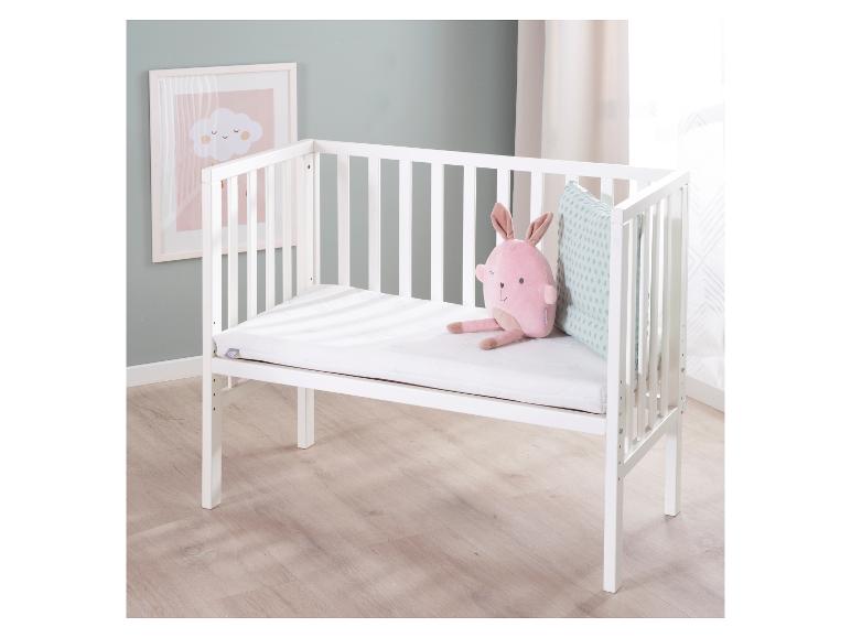 Wit babybedje met een roze knuffel en een kussen, naast een wolkenafbeelding.