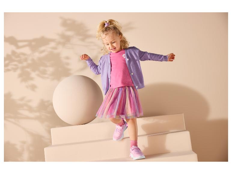 Lachend meisje in een roze top, paars vest en kleurrijke rok, met roze sneakers.