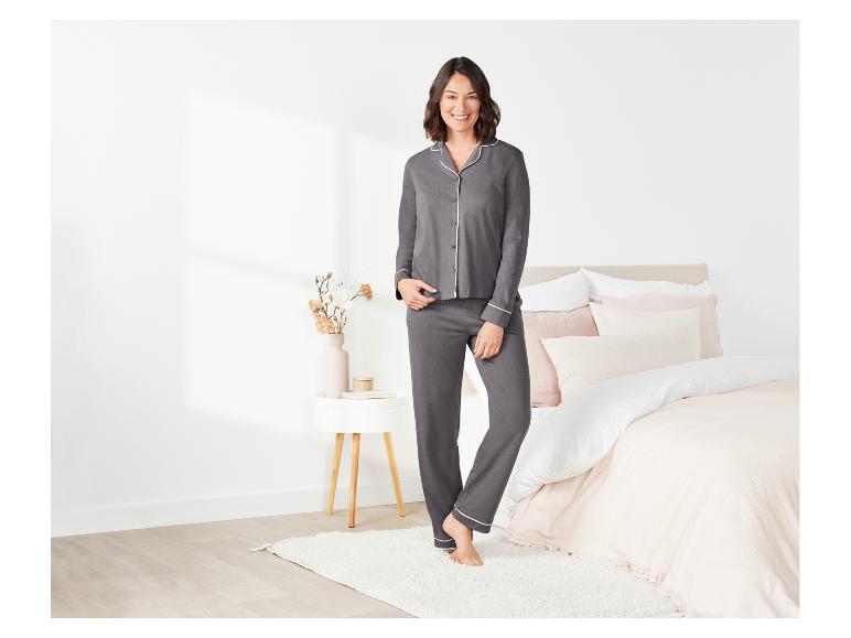 Een vrouw in een grijze pyjama met witte piping staat voor een bed.