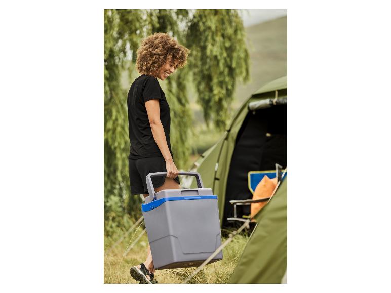 Lachende vrouw draagt een grijze koelbox naast een kampeertent.
