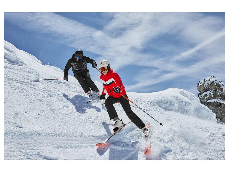 Twee skiërs in skijassen en skibroeken, dalend van een besneeuwde helling.