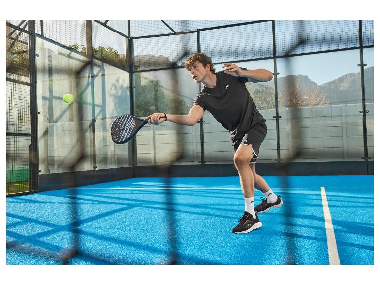Man speelt padel op blauwe baan, draagt zwart T-shirt en korte broek, met racket in hand.