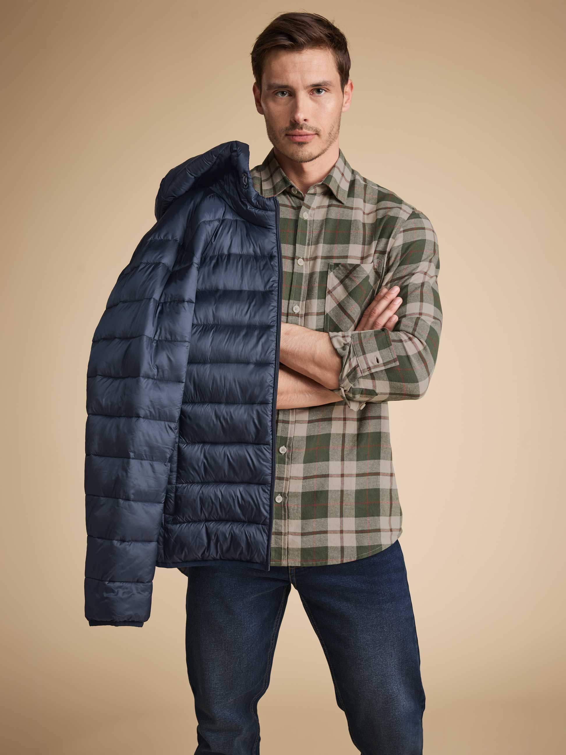Man in een groen geruit overhemd en jeans, met een blauwe gewatteerde jas over zijn schouder.