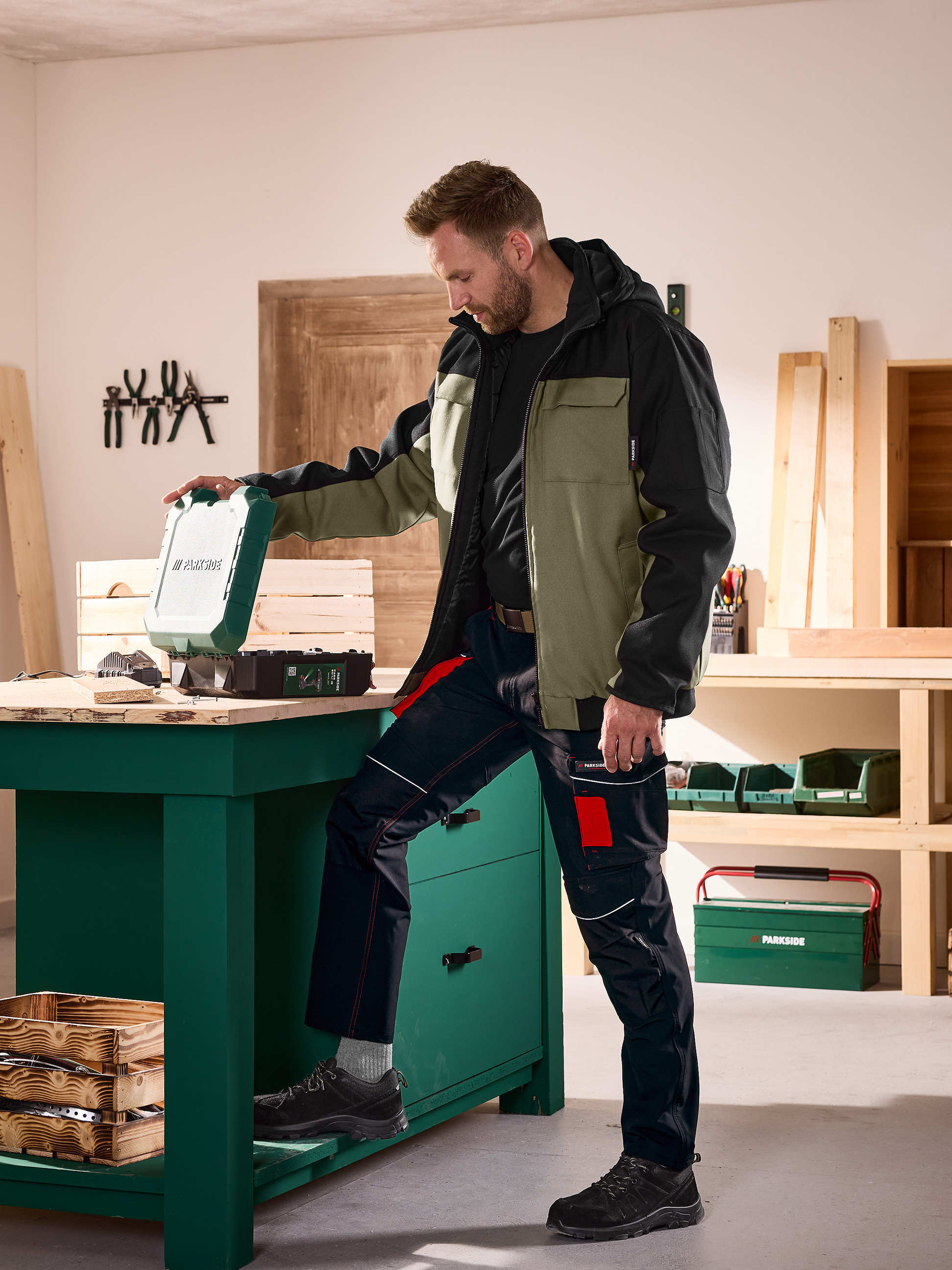 Man in werkjas en -broek opent een gereedschapskoffer op een werkbank in een werkplaats.