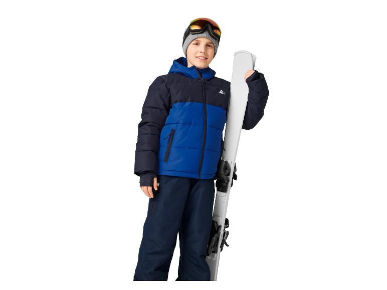 Een lachende jongen in een blauw-zwarte ski-jas, broek en skibril, met ski's.