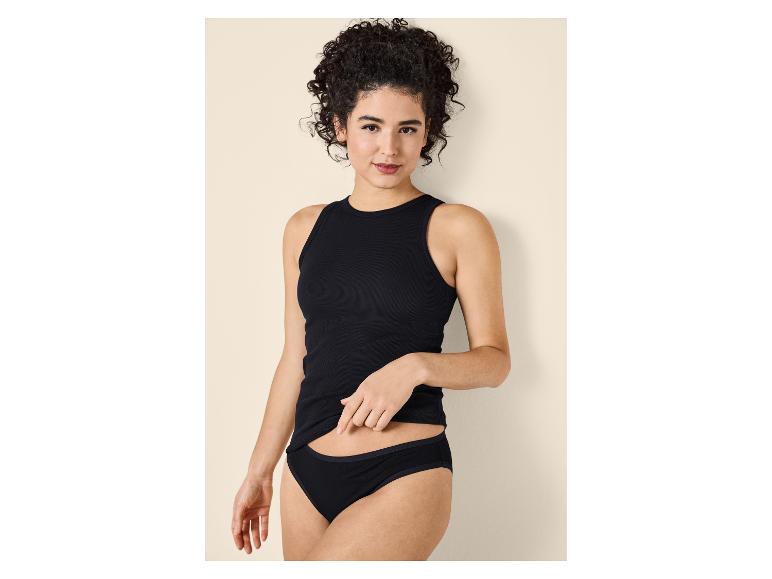 Vrouw met krullend haar in een zwart hemdje en slip, poserend tegen een beige achtergrond.
