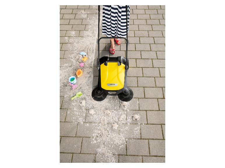 Vrouw in gestreepte jurk en rode sandalen duwt gele veegmachine over bestrating, zand vegend.