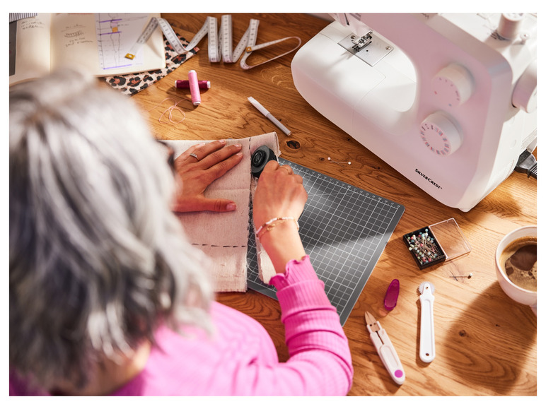 Een vrouw die met een rolmes op stof werkt, met een naaimachine en andere naaigerei ernaast