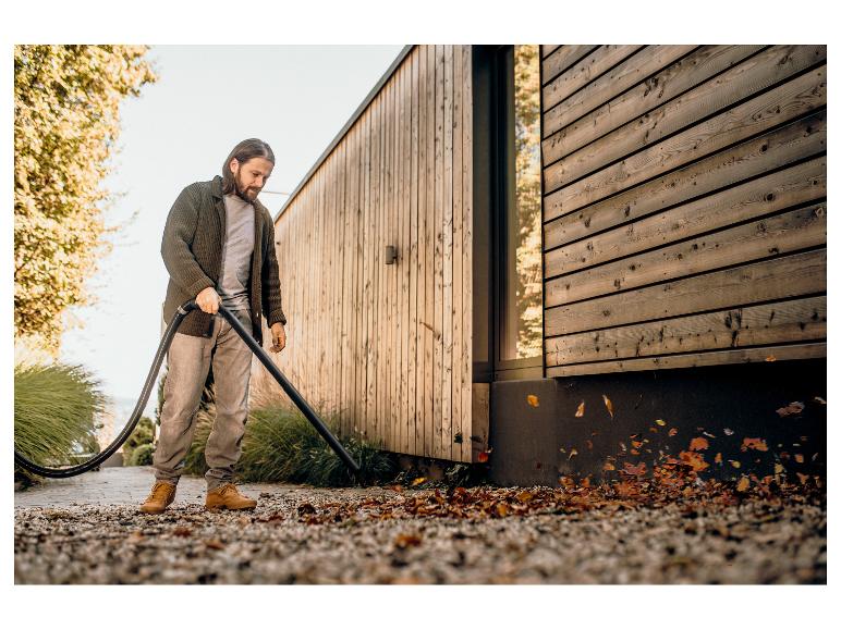 Man met bladblazer ruimt bladeren op bij houten huis.
