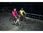 Twee mensen in reflecterende fietsjacks rijden 's nachts op elektrische fietsen langs een rivier.