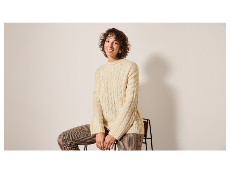 Een vrouw draagt een crèmekleurige kabeltrui en een broek met ruitjespatroon.