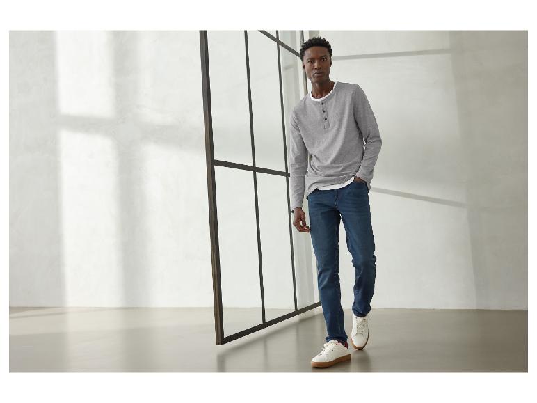 Man in grijs shirt met lange mouwen, blauwe jeans en witte sneakers.