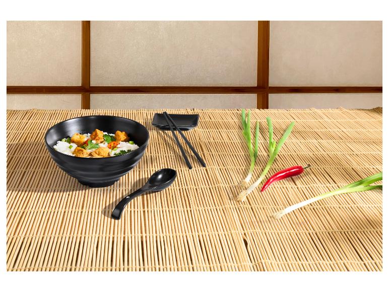 Zwarte kom met Aziatisch eten, eetstokjes, lepel, lente-uitjes en chili op een bamboemat.