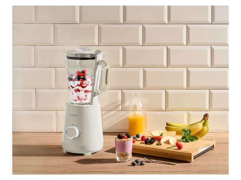 Een blender gevuld met bessen en yoghurt, naast een glas smoothie en vers fruit.