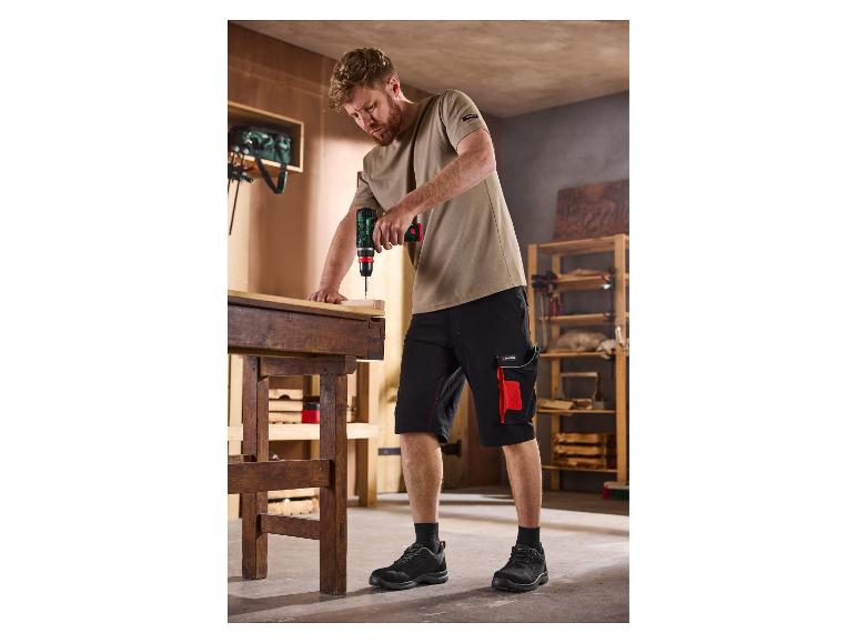 Man in werkshorts en T-shirt gebruikt een accuboormachine op een houten werkbank.