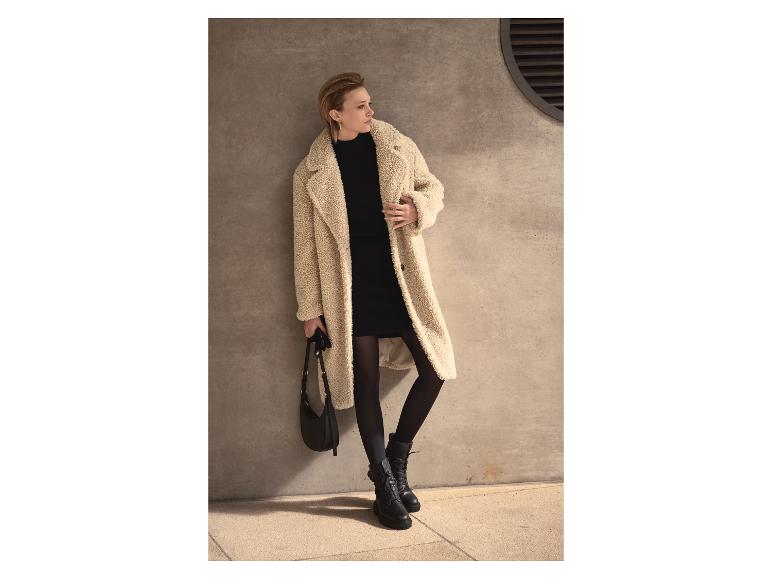 Vrouw in een crèmekleurige teddyjas, zwarte jurk, panty en laarzen, met een zwarte tas.