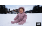 Lachende baby in roze sneeuwpak zittend in de sneeuw, met een huis en bomen op de achtergrond.