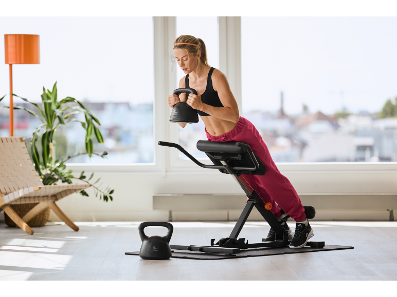 Vrouw traint met kettlebell op een rugtrainer in een lichte kamer.