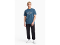 Man in een blauw T-shirt met ruitpatroon, zwarte joggingbroek en witte sneakers.