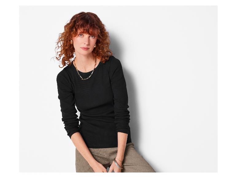 Vrouw met rood haar, draagt een zwart shirt met lange mouwen en een bruine broek.