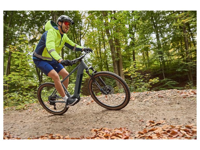 Man in neongele jas en blauwe korte broek fietst op een e-bike op een bospad.