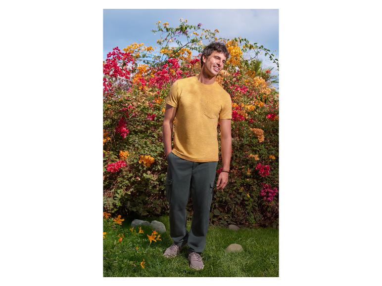 Man in geel T-shirt en donkergroene broek poseert voor bloemen.