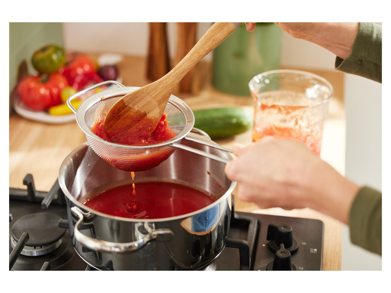 Een hand giet gepureerde tomaten uit een zeef in een pan op het fornuis.