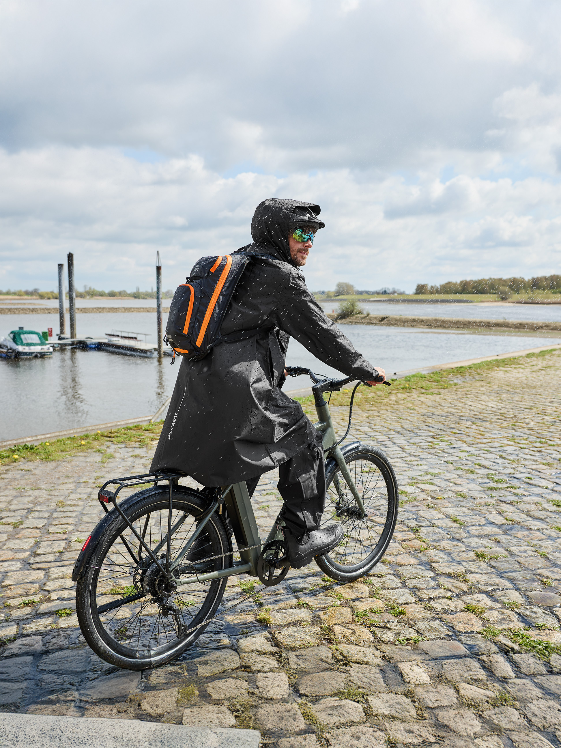 Man in regenjas met rugzak fietst in de regen langs de rivier.