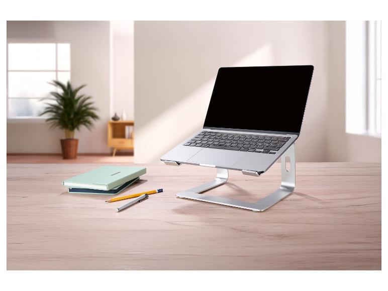 Zilveren laptopstandaard met een laptop, notitieboekjes en pennen op een houten tafel.