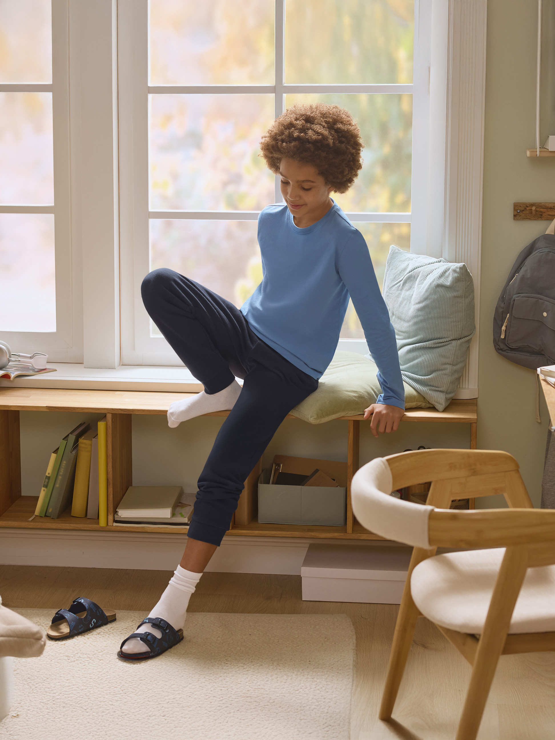 Jongen in blauwe pyjama zit op vensterbank met boeken en kussens.