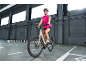 Lachende vrouw in roze sportkleding en helm, rijdend op een grijze elektrische fiets in een magazijn.
