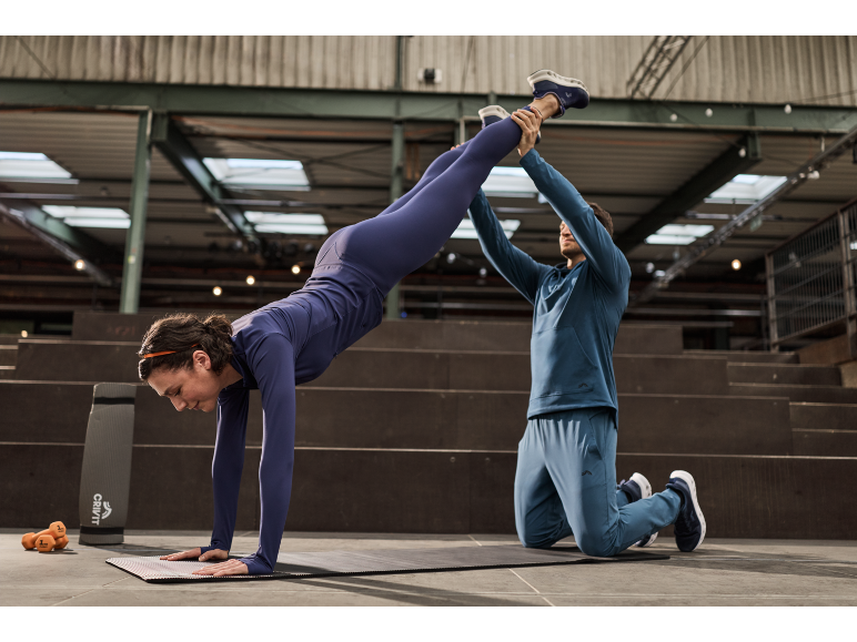 Vrouw in blauwe sportkleding doet een handstand op een mat met hulp van een trainer.