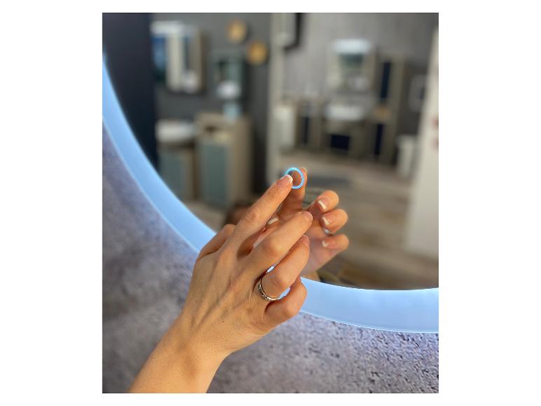 Vrouwenhanden raken een rond verlichte spiegel aan, die een moderne badkamer weerspiegelt.