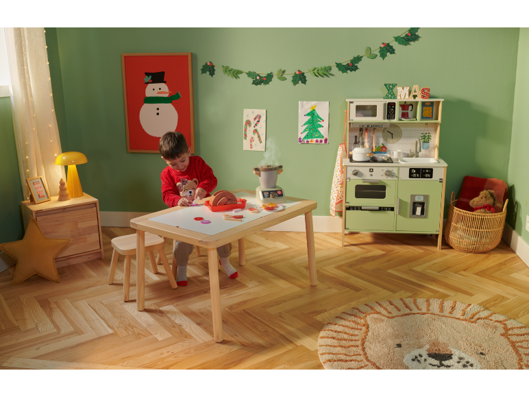 Kind speelt met een houten speelkeuken en speelgoedeten in een kerstversierde kamer.