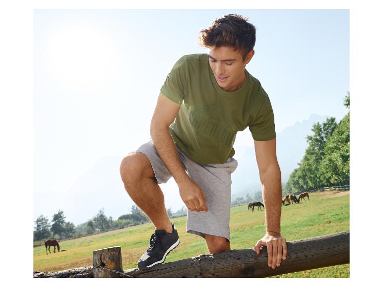 Man in groene T-shirt en grijze shorts, staat op een hek in een weiland met paarden.