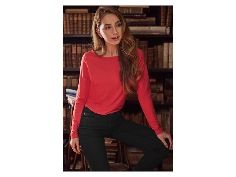 Vrouw in een rood shirt met lange mouwen en zwarte jeans, leunend op een stoel voor een boekenplank.