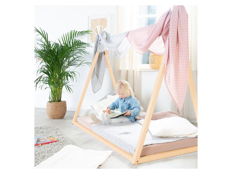 Een kind leest een boek in een houten Montessori-bed, versierd met roze en grijze stoffen.