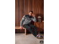 Man in een grijs patroonvest en donkere jeans, zittend op een houten bank.