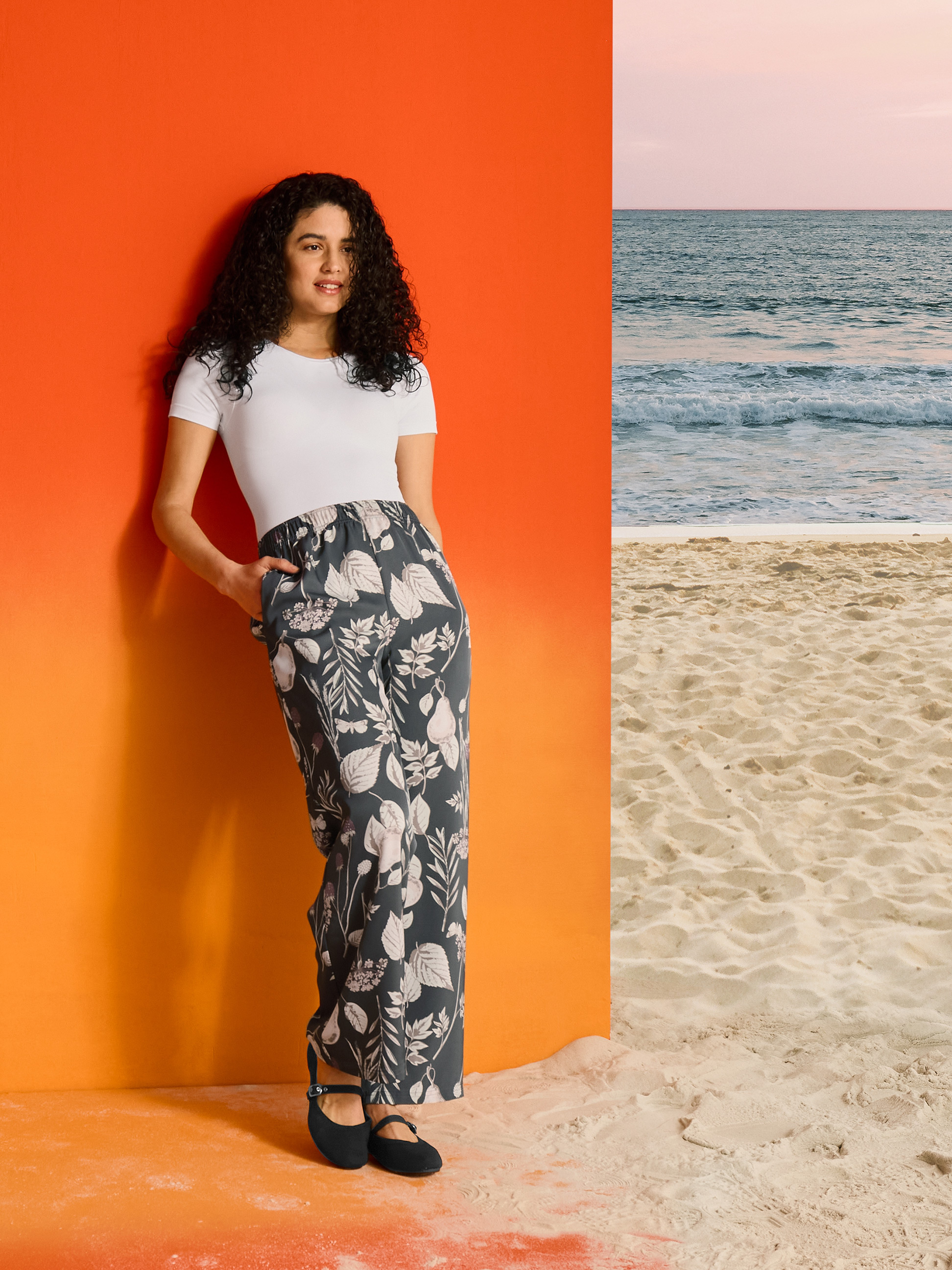 Vrouw in bloemenbroek en wit T-shirt, tegen oranje achtergrond en strand.