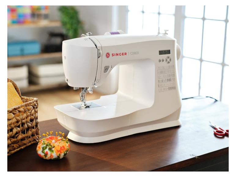 Witte Singer naaimachine op een houten tafel, met een mandje benodigdheden en een speldenkussen.