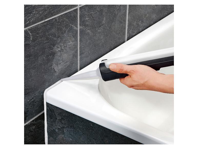 Een hand brengt witte kit aan op de rand van een badkuip naast donkere tegels.