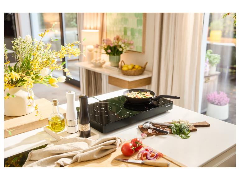 Een zwarte inductiekookplaat met een koekenpan die groenten kookt, omringd door verse ingrediënten.