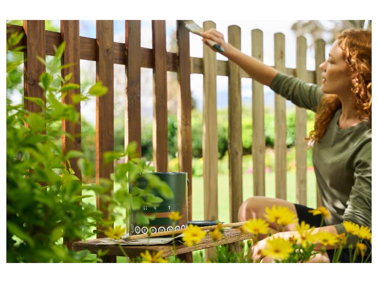 Vrouw schildert houten hek met groene verf, kwast en gele bloemen.